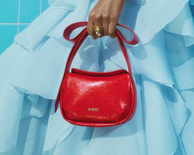 Hand holding a red Mimco handbag with a curved top handle, set against a light blue ruffled dress.
