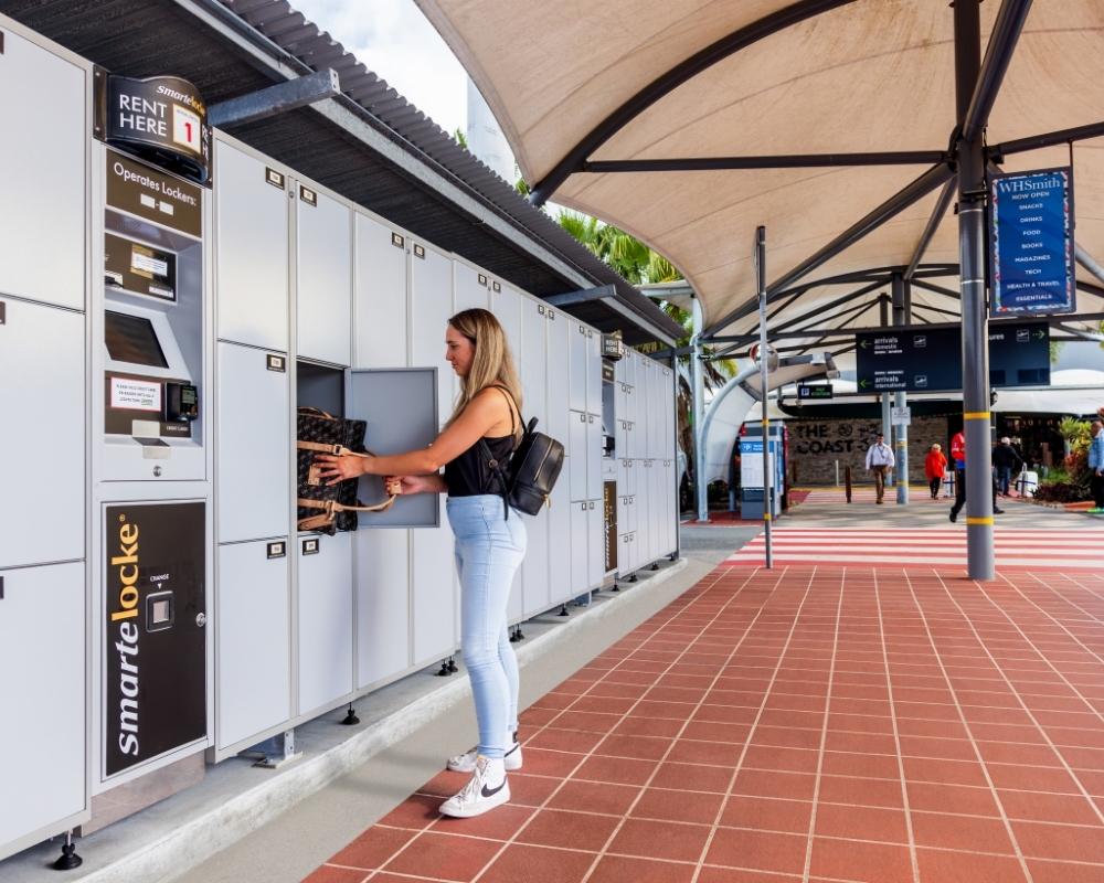 A woman uses a Smarte Carte luggage locker system at an outdoor terminal area. She is placing or retrieving a bag from a secure compartment under a shaded walkway with signs and people in the background.