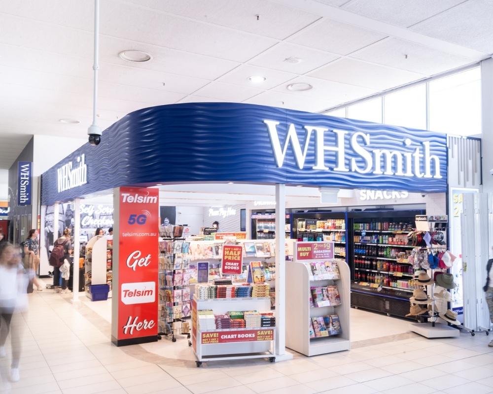 The exterior of a WHSmith store at an airport, featuring bright signage, promotional displays, and shelves stocked with books, snacks, drinks, and travel essentials. Passengers walk by in the terminal corridor.