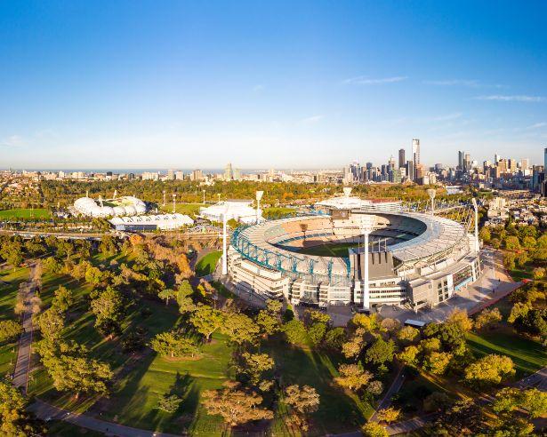 Aerial view of the Melbourne Cricket Ground (MCG) on a sunny day, surrounded by the parklands of Yarra Park with the Melbourne city skyline behind.