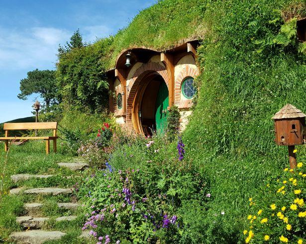 A Hobbit-style home built into a lush green hillside, with a round wooden doorway surrounded by colourful flowers and a garden path.