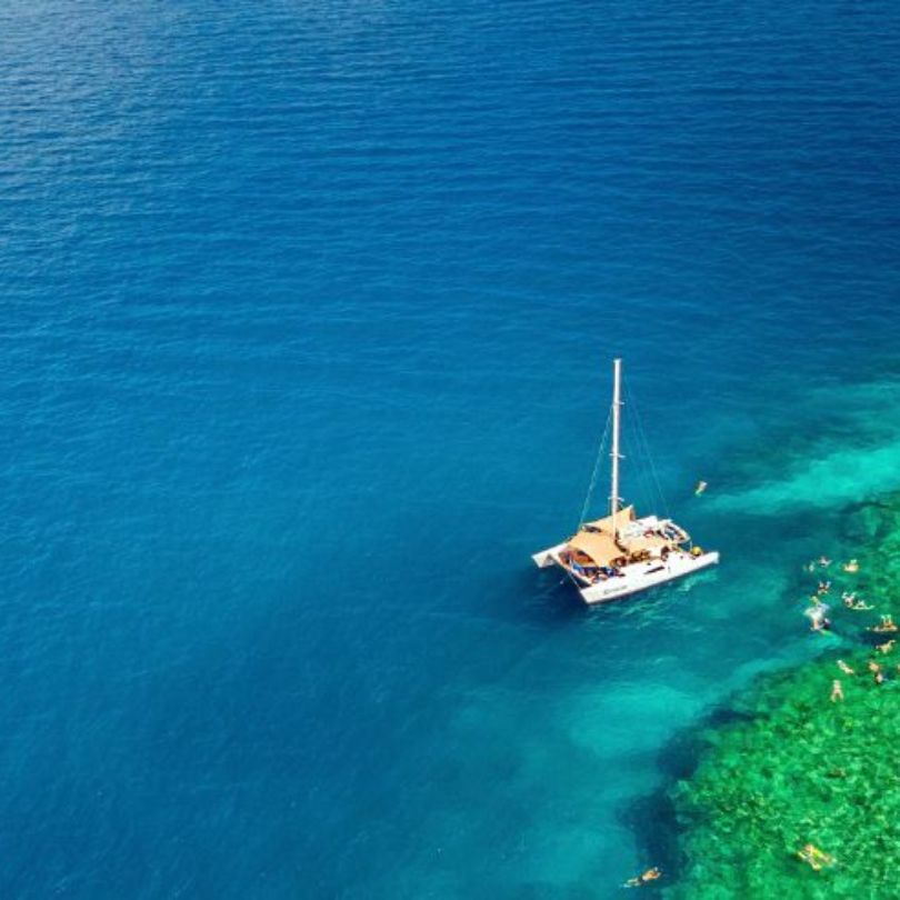 An aerial view of a boat floating on deep blue ocean water beside a bright turquoise reef where people are swimming and snorkelling.