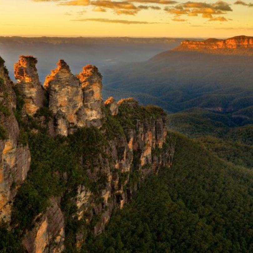 The Three Sisters rock formation in the Blue Mountains, surrounded by forested valleys and sandstone cliffs at sunset.