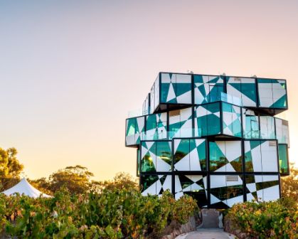 Picture of The d’Arenberg Cube in McLaren Vale, South Australia, a striking multi-storey building with an abstract, cube-like design made of glass panels in geometric patterns, surrounded by vineyards at sunset.