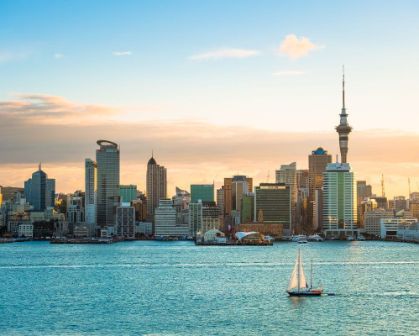 Picture of A scenic view of Auckland’s skyline at sunset, featuring the Sky Tower and waterfront buildings. A sailboat glides across the calm harbour waters in the foreground.