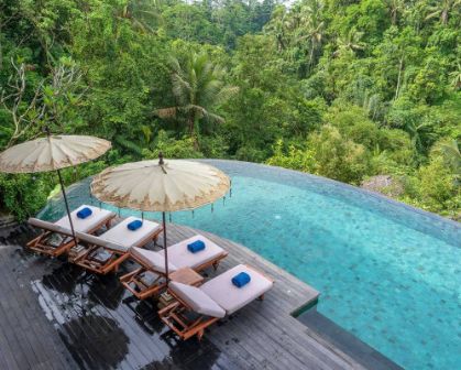 Picture of A tranquil infinity pool overlooking lush jungle in Bali, with four sun loungers topped with rolled blue towels and shaded by traditional Balinese umbrellas on a wooden deck.