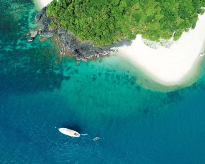 Picture of Aerial view of a tropical island with lush green foliage, white sandy beach, and clear turquoise water. A boat floats just offshore, surrounded by vibrant blue ocean and coral reefs.