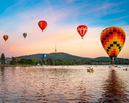 Picture of Colourful hot air balloons float above a lake at sunrise in Canberra, Australia. Reflections dance on the water as kayakers paddle below, with Telstra Tower visible atop the hill in the background.