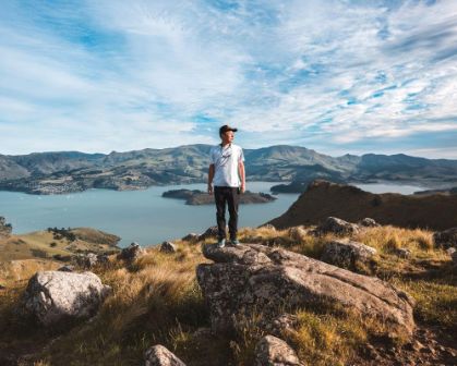 Picture of A man stands on a rocky hilltop with panoramic views of Christchurch’s Lyttelton Harbour in New Zealand. Surrounded by golden grass and rugged terrain, he overlooks the deep blue bay and rolling hills under a dynamic sky.