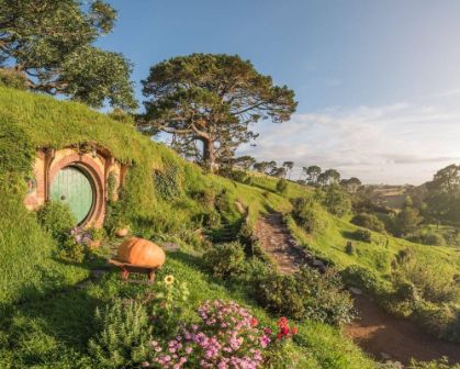 Picture of A picturesque view of Hobbiton in Matamata, New Zealand, featuring a round green hobbit door nestled in a hillside, surrounded by flowers and a winding garden path under golden morning light.