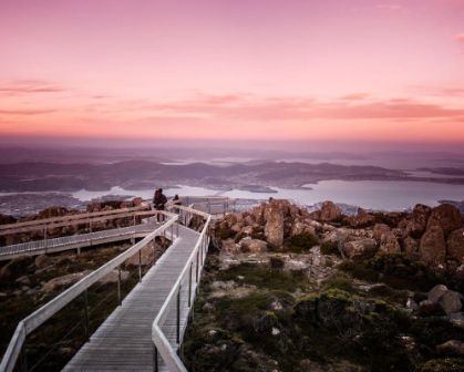 Picture of Elevated walkway on kunanyi / Mount Wellington overlooking Hobart, Tasmania, at sunset. The sky glows pink and purple as visitors take in panoramic views of the city, harbour, and surrounding landscape.