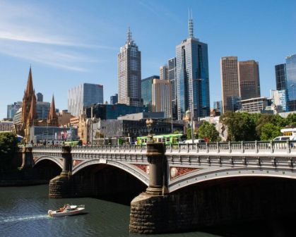 Picture of Scenic view of Melbourne’s city skyline with a mix of historic and modern buildings, including St Paul’s Cathedral. The Princes Bridge crosses the Yarra River, with a boat cruising beneath and a tram crossing above.
