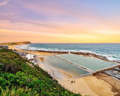 Picture of Coastal view of Newcastle Ocean Baths in New South Wales at sunset, with swimmers enjoying the tidal pool beside the sandy beach and waves rolling in from the ocean under a pastel-coloured sky.