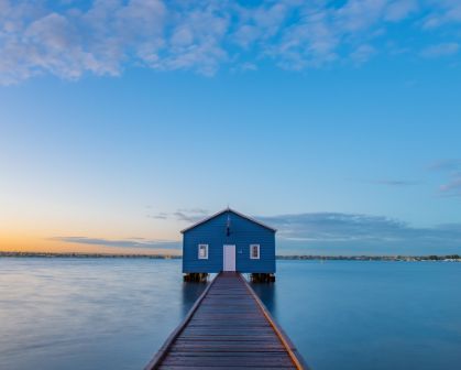 Picture of The iconic blue Crawley Edge Boatshed in Perth, Western Australia, sits at the end of a wooden jetty on the calm waters of the Swan River at sunrise, under a clear blue sky.