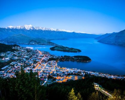 Picture of A twilight view of Queenstown, New Zealand, with the town’s lights glowing beside Lake Wakatipu. Snow-capped mountains of The Remarkables range rise in the distance under a deep blue sky.