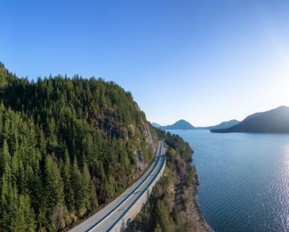 Picture of Aerial view of the Sea to Sky Highway near Vancouver, curving between lush green forest and the sparkling waters of Howe Sound, with mountains on the horizon.