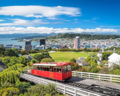 Picture of Iconic red Wellington Cable Car travelling uphill with panoramic views over Wellington city, lush gardens, and the harbour on a clear day.