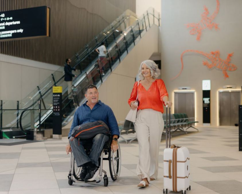  A man in a wheelchair and a woman with a suitcase walk together through a spacious airport terminal. Behind them are escalators, lifts, seating, and a red gecko wall mural, contributing to a bright, modern atmosphere.