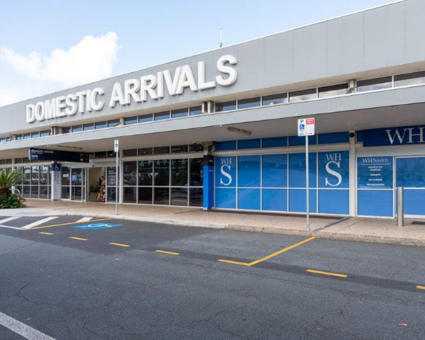 Domestic Arrivals entrance at Gold Coast Airport, featuring large white signage, glass doors, blue WHSmith window panels, and designated parking bays in the foreground.
