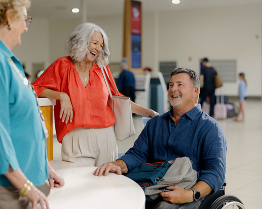 A man in a wheelchair smiles while speaking with two women at an airport assistance desk. One woman wears a red blouse and the other is in a teal uniform. The terminal background shows check-in kiosks and travellers with luggage.