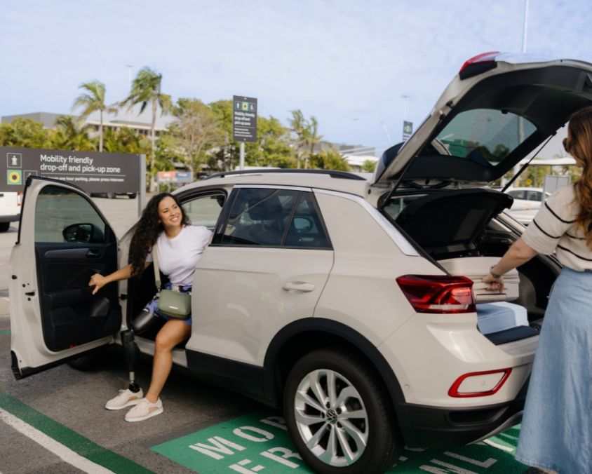 A woman with a prosthetic leg smiles as she exits a car parked in an accessible parking bay at the airport. Another person stands at the open boot, with palm trees and signage visible in the background.