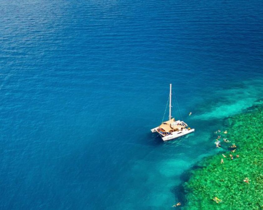 An aerial view of a boat floating on deep blue ocean water beside a bright turquoise reef where people are swimming and snorkelling.