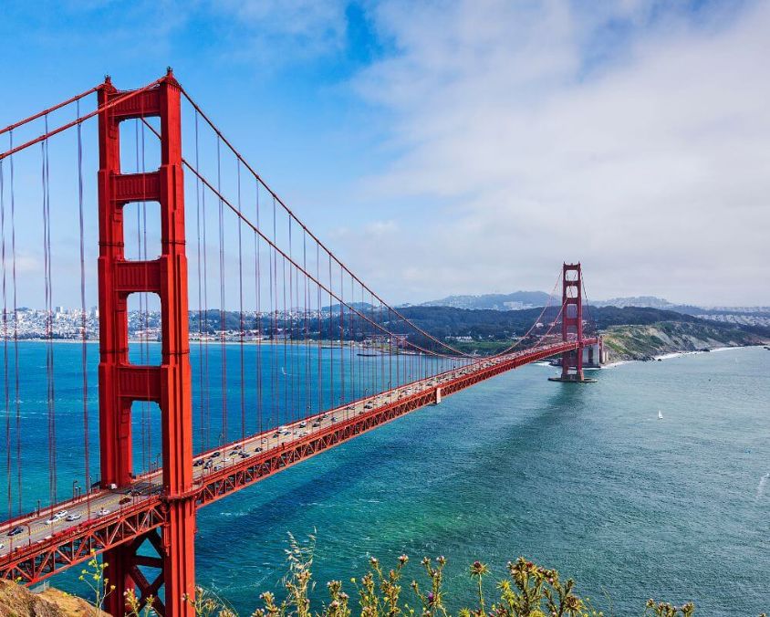 The Golden Gate Bridge in San Francisco, California, stretches across the bay under a partly cloudy sky. The iconic red suspension bridge is surrounded by blue-green waters and rolling hills.