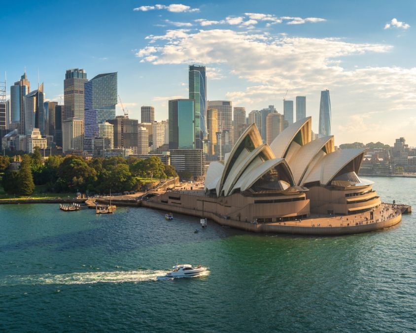 Aerial view of the Sydney Opera House at sunset, surrounded by Sydney Harbour and the city skyline. A white boat cruises across the sparkling water in the foreground.