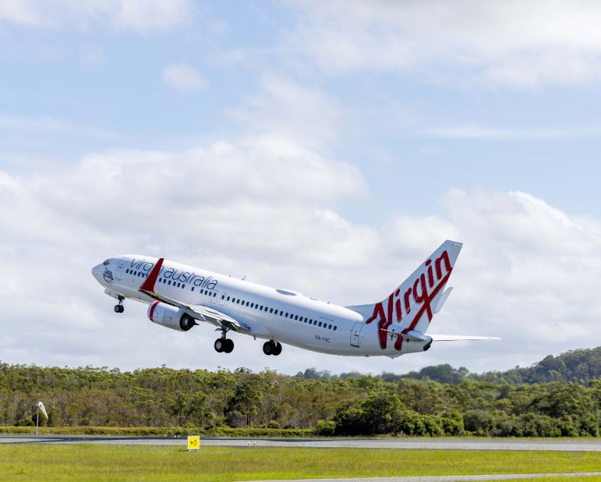A Virgin Australia airplane, a Boeing 737, is captured mid-takeoff, ascending from the runway. The aircraft's white fuselage features the airline's red branding, with "Virgin" prominently displayed on the tail. The background consists of a partly cloudy sky and lush green trees near the airport. The landing gear is retracting as the plane gains altitude.