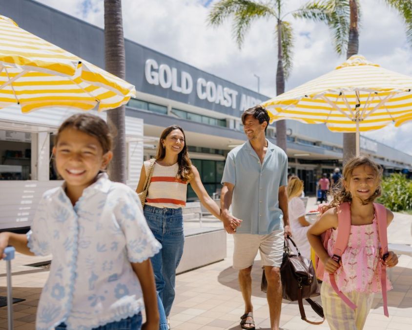 "A smiling family of four walks outside Gold Coast Airport on a sunny day. The parents, a woman in a striped top and jeans and a man in a light blue shirt and shorts, hold hands while carrying travel bags. Two young girls, one wearing a pink backpack and the other pulling a suitcase, walk ahead excitedly. Yellow and white striped umbrellas and palm trees line the walkway, creating a bright and welcoming atmosphere.