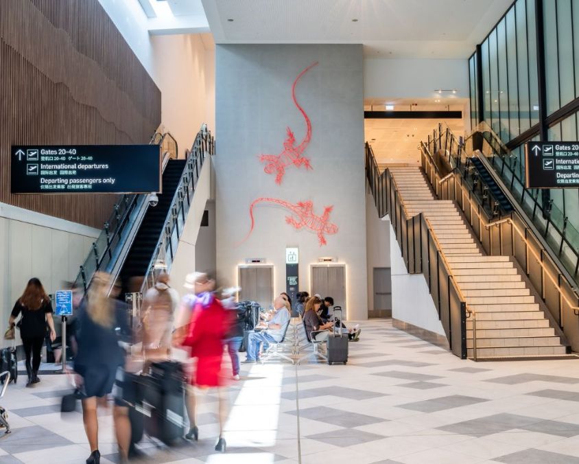 Passengers move through a modern airport terminal with escalators, a staircase, and red gecko-inspired wall art above a lift.