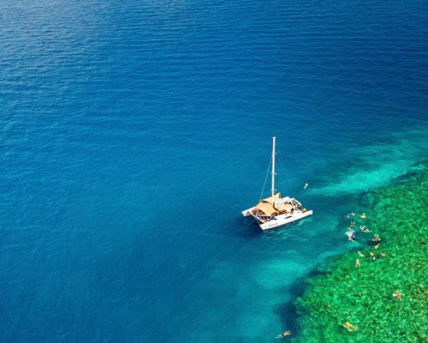 Aerial view of a white catamaran anchored beside a vibrant coral reef, with people snorkelling in clear turquoise water surrounded by deep blue ocean.