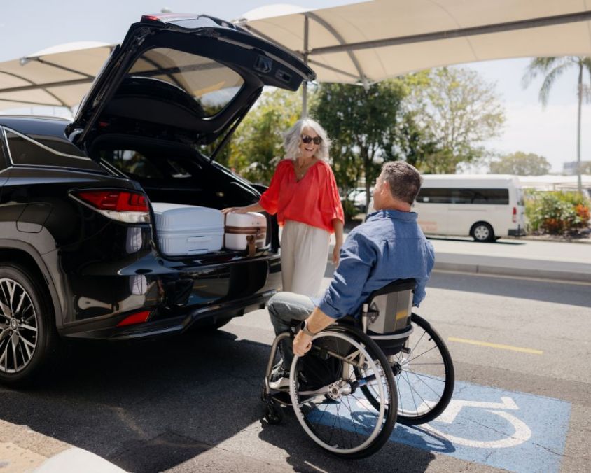A man using a wheelchair and a woman retrieve luggage out of the boot of a car at Gold Coast Airport, with a shade sail visible behind them.