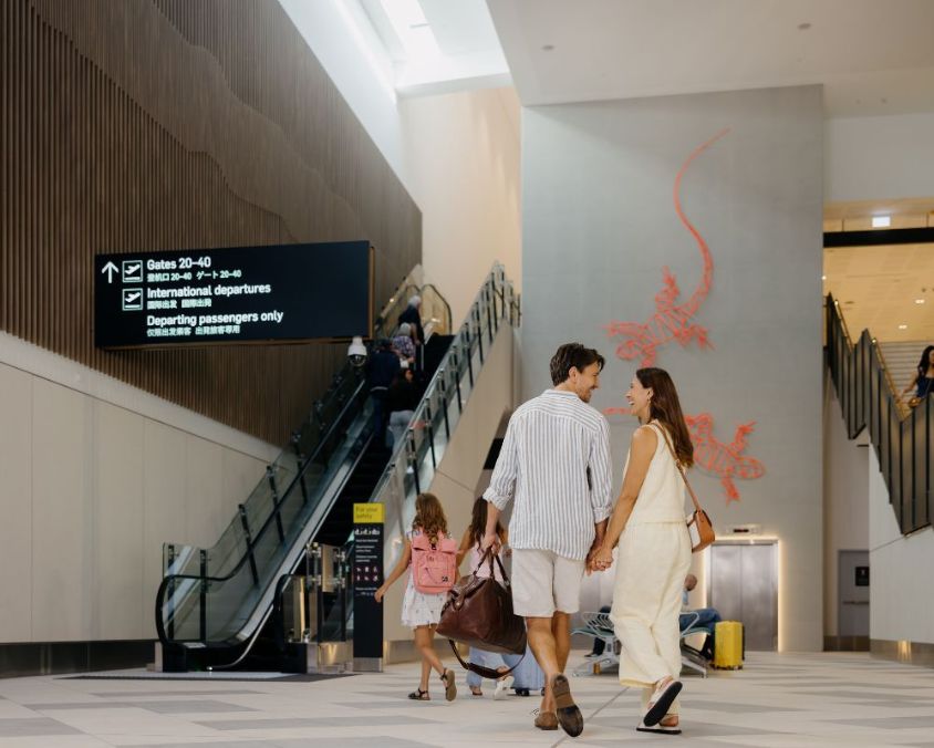 A family walks through Gold Coast Airport past the red gecko wall art and escalators, making their way towards international departures.