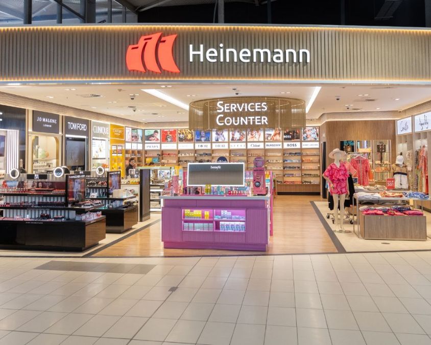 Front view of the Heinemann retail store at an airport, showcasing a well-lit interior with beauty counters for brands like MAC, Tom Ford, Clinique, and Benefit. The Services Counter is centrally located, and the right side of the store displays mannequins dressed in bright resort wear and summer accessories. The store has clear signage and a modern, inviting design.