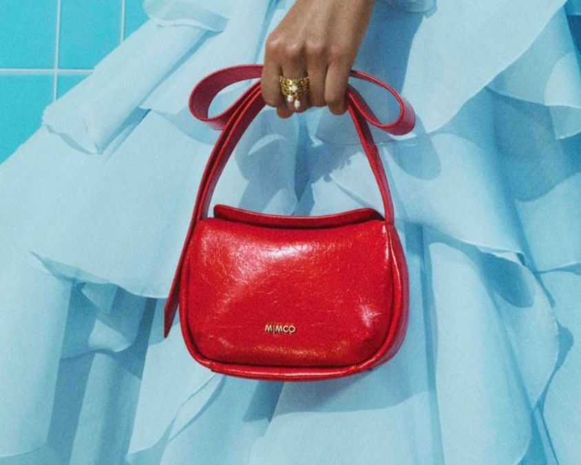 Hand holding a red Mimco handbag with a curved top handle, set against a light blue ruffled dress.
