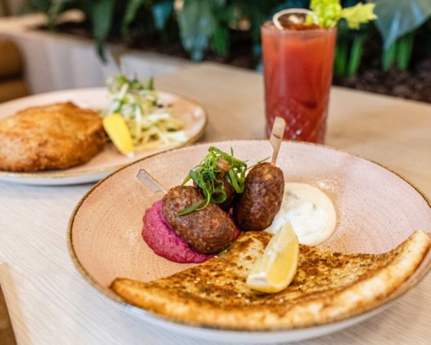 Plate of grilled kofta skewers served with flatbread, dips, lemon wedge, and a Bloody Mary drink on a table, with another plated meal in the background.