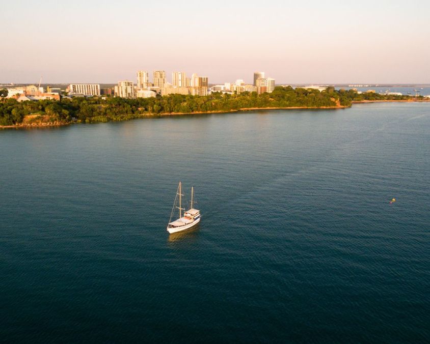 A sailboat glides across calm blue waters at sunset, with the Darwin city skyline and lush green coastline in the background.