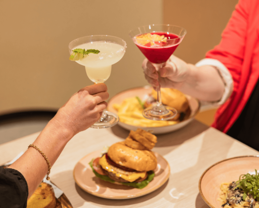 Two people clinking cocktail glasses over a table with plates of burgers, fries, and pasta at a restaurant.