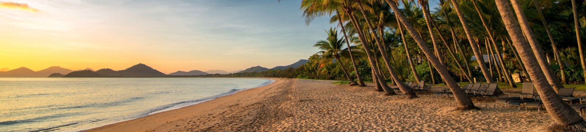 Palm-fringed sandy beach in Cairns at sunrise, with calm ocean waves and distant mountains under a golden sky.