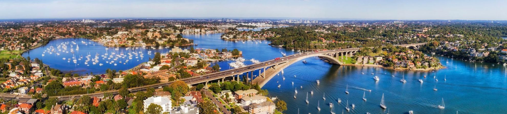 Aerial panoramic view of Western Sydney, showing a wide river dotted with boats, a large bridge crossing the water and surrounding residential suburbs.