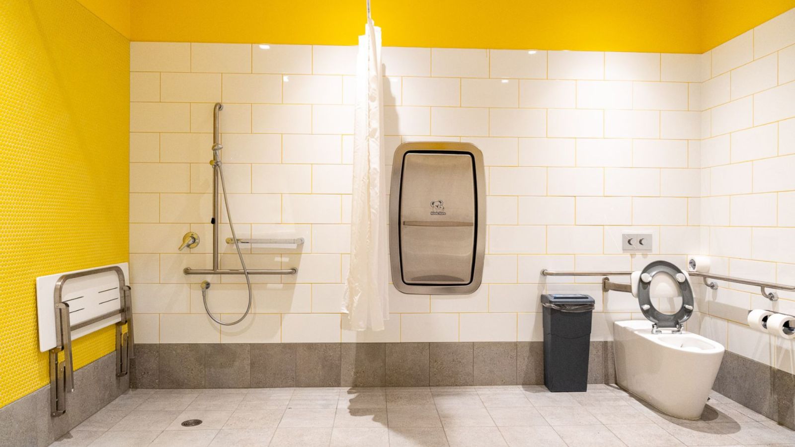 A brightly lit accessible bathroom with yellow and white tiled walls, featuring a roll-in shower with fold-down seat and grab rail, a toilet with side grab bars, a sanitary bin, and a curtain for privacy.