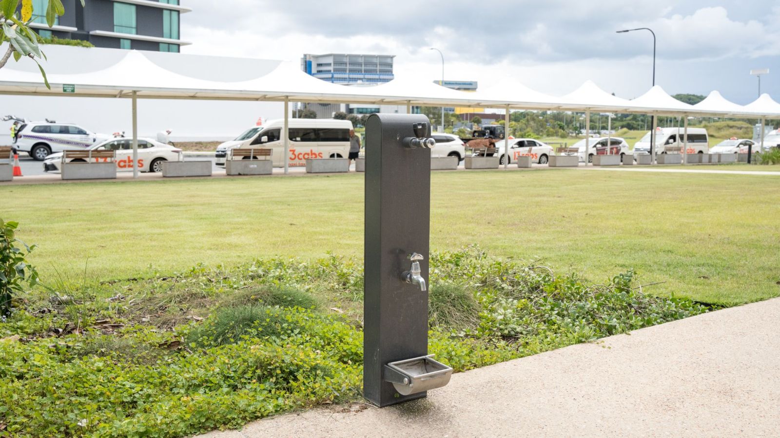 An outdoor water fountain with a bottle refill tap and dog bowl feature, located on a grassed area near an airport pickup zone. Taxis and white shade structures are visible in the background.