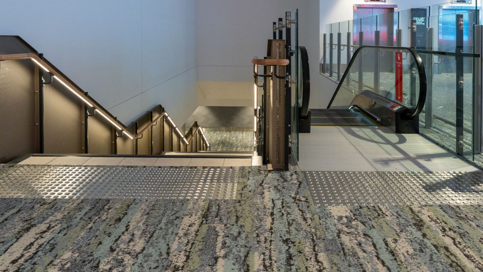 Airport interior featuring a carpeted area leading to a stairway and an escalator. Tactile ground surface indicators are installed at the top of the stairs for accessibility, with soft lighting along the stair rails.