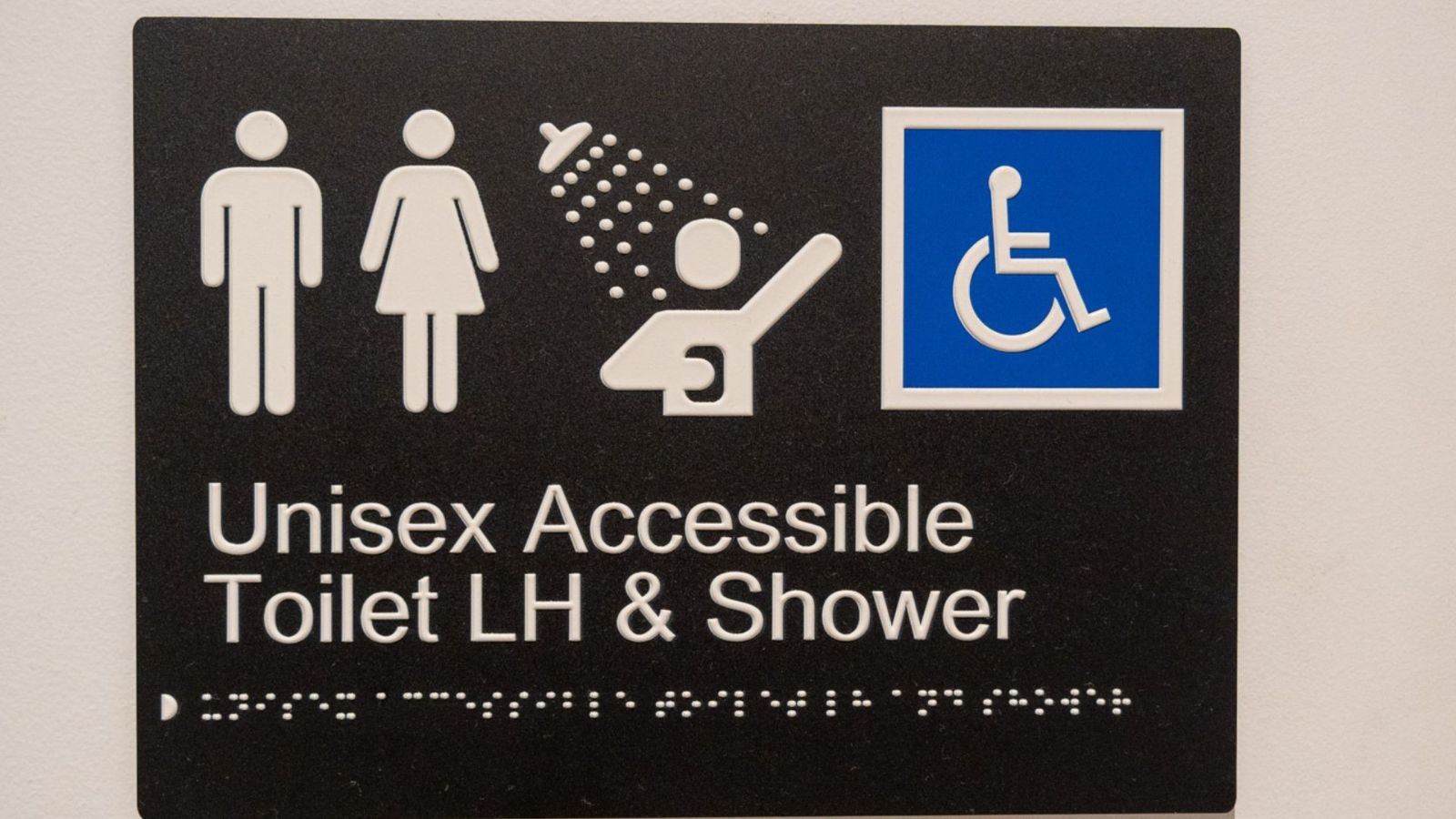 Sign for a Unisex Accessible Toilet with Left-Hand (LH) transfer and shower, featuring icons for male, female, accessibility, and a shower with a seat. Braille is included beneath the text.