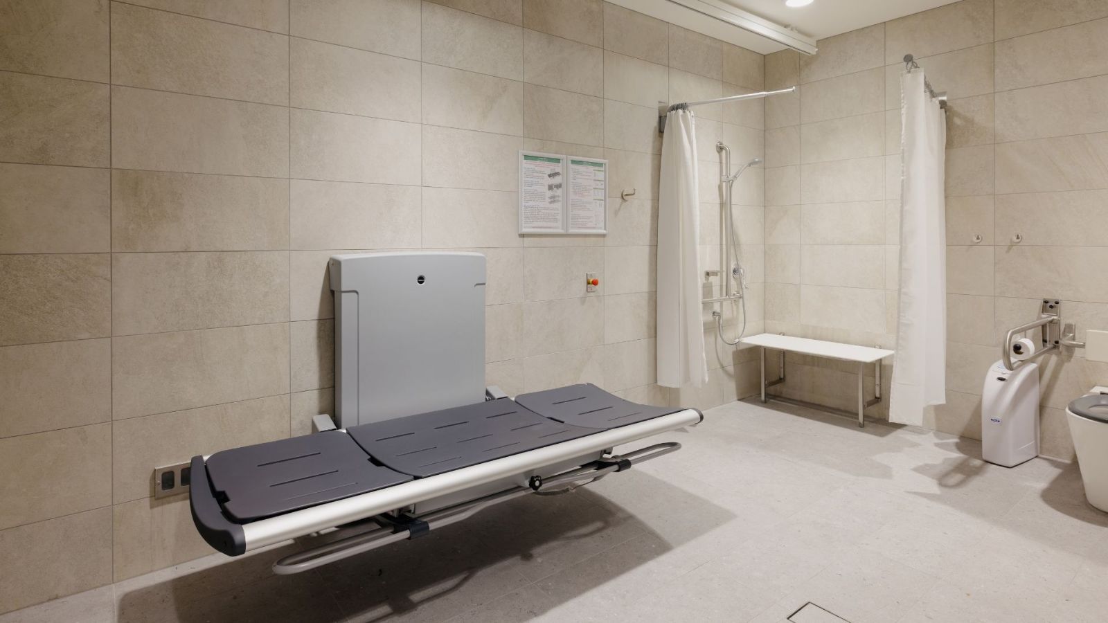Inside an airport’s Changing Places facility, featuring a height-adjustable adult-sized change table, a ceiling track hoist, a centrally located peninsular toilet, a privacy screen, grab rails, sensor lighting, and an accessible shower. The space is designed to accommodate a traveller with a disability and up to two carers.