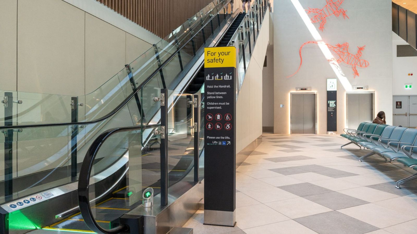 Airport interior with an escalator, elevator access, and seating area. Safety signage is displayed near the escalator, and a red wall mural of a kangaroo adds a local touch. The space is well-lit with natural light from above.