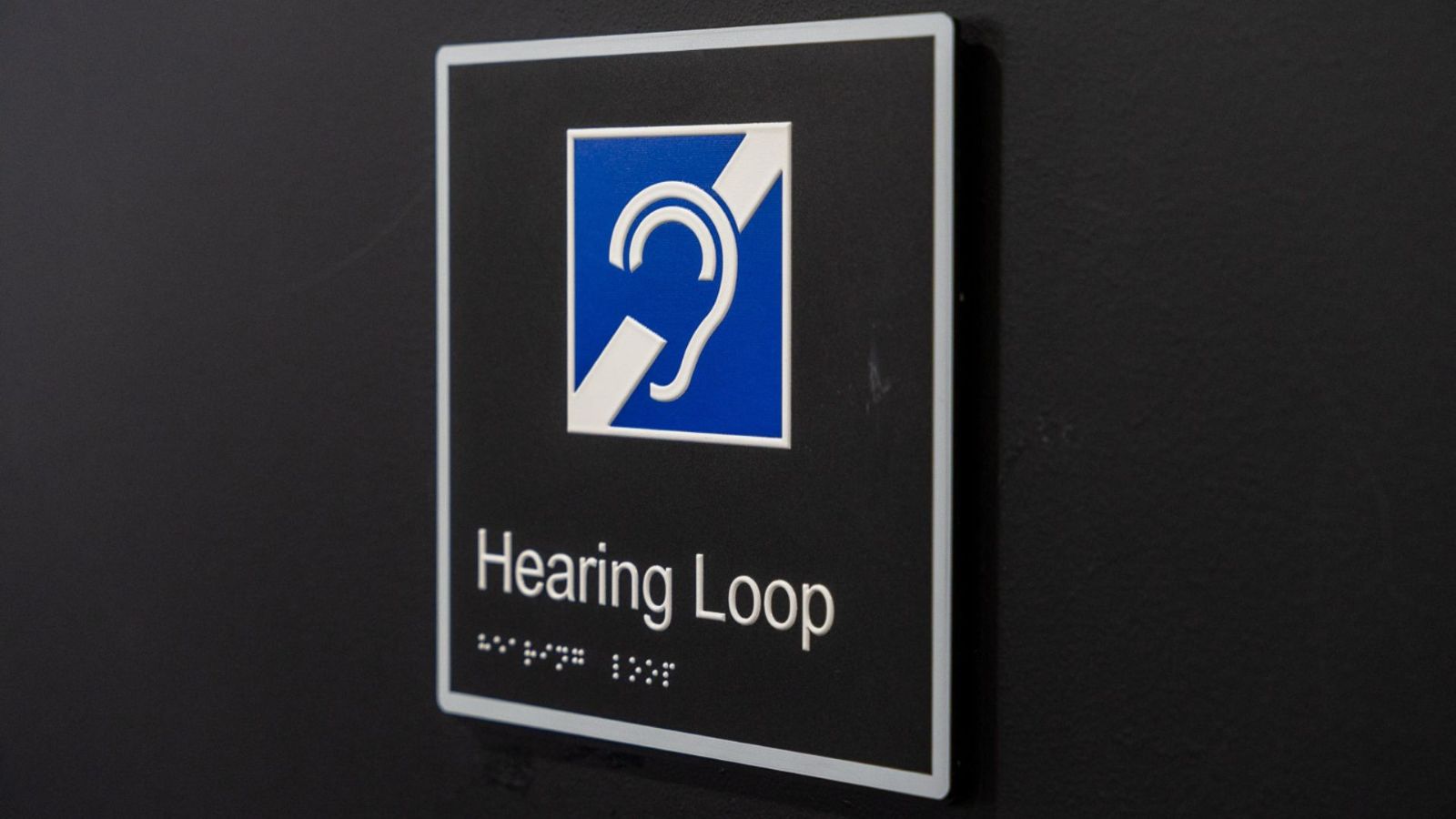 A black sign indicating the availability of a Hearing Loop system, featuring a blue and white ear symbol with a diagonal slash and braille text below the words "Hearing Loop."