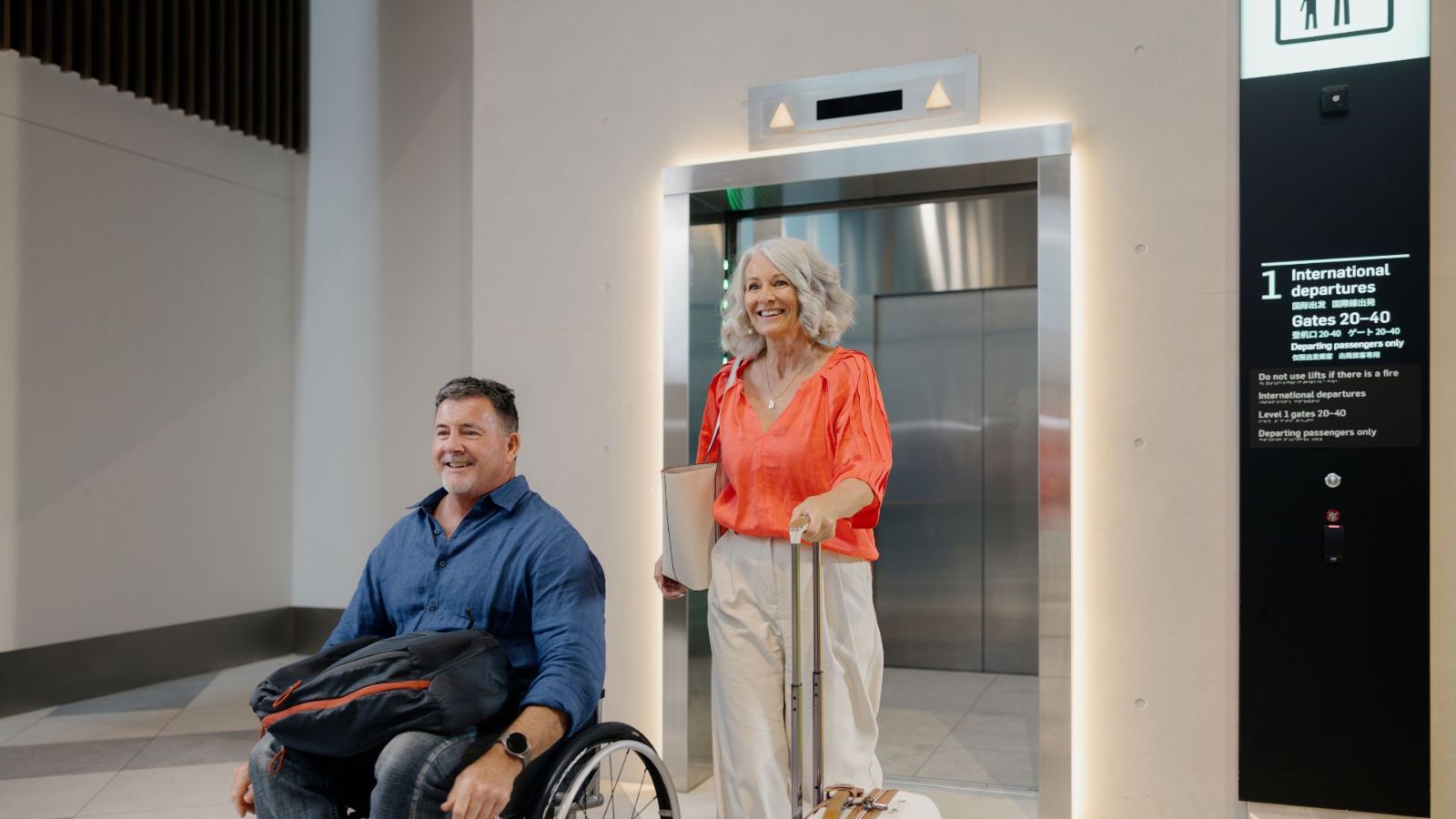 A man in a wheelchair and a woman with luggage exit an airport lift together. The space is modern and well-lit, with accessible signage visible beside the elevator.