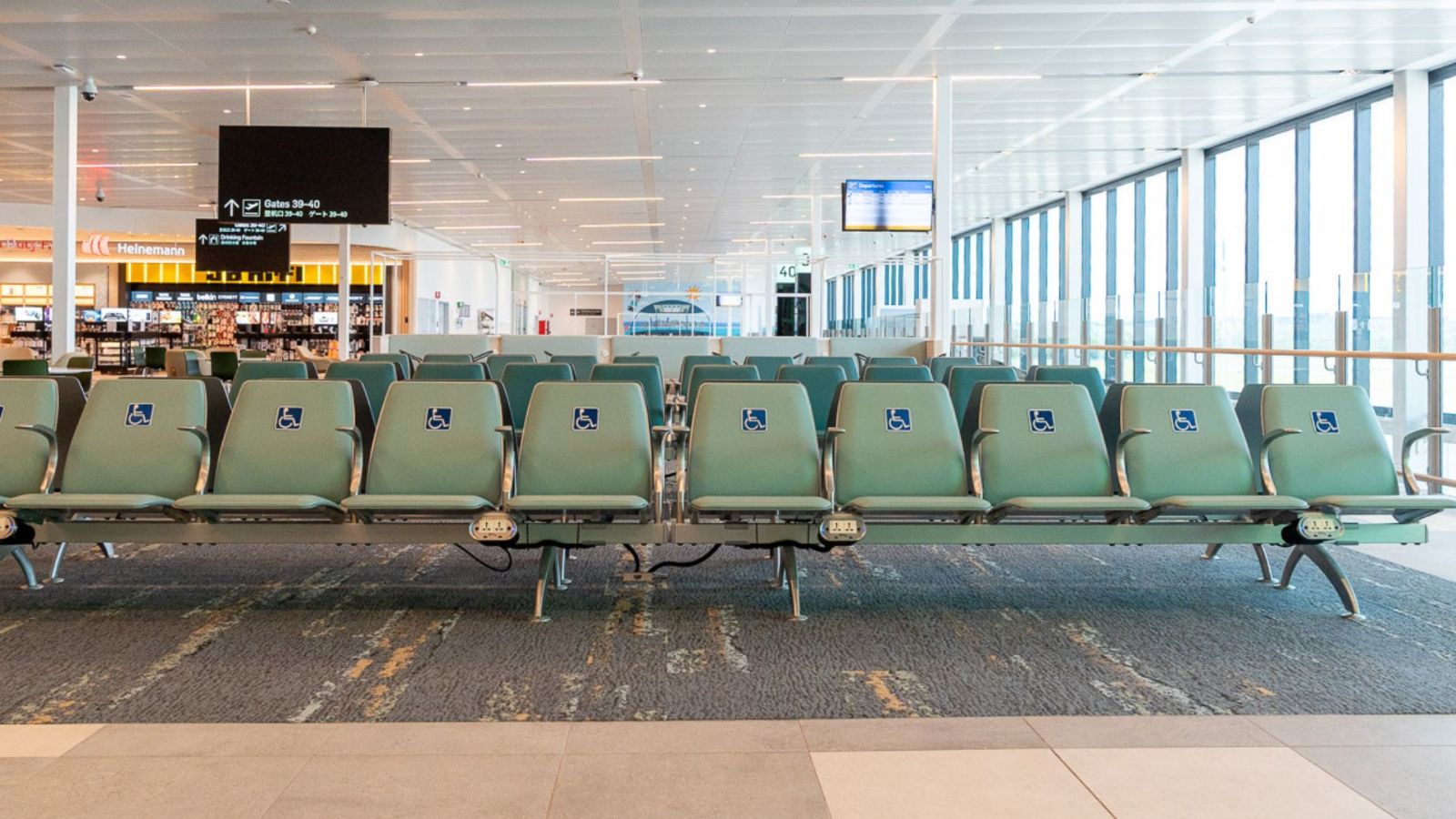 A row of green airport terminal seating with designated priority seats marked by blue accessibility symbols. Large windows line the right side, allowing natural light into the spacious boarding area.
